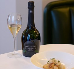 Flute of champagen on the left, with the bottle of Laurent Perrier Grand Siecle to the right. In front is a dish.