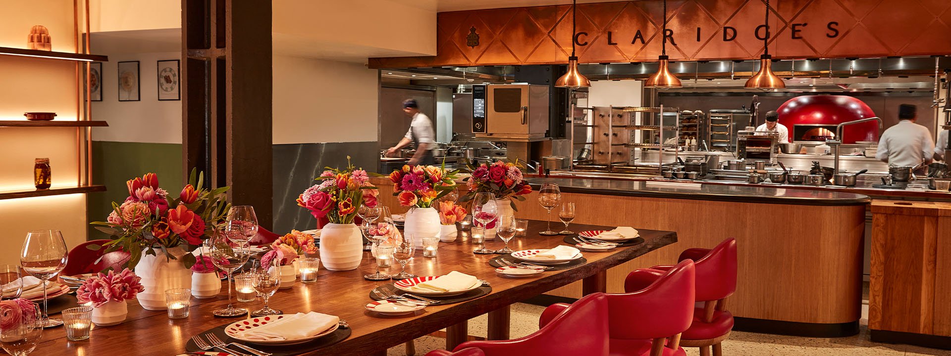 A lively chef’s table setup with red chairs and floral centrepieces facing an open kitchen at Claridge’s, where chefs are seen preparing food under copper lighting.