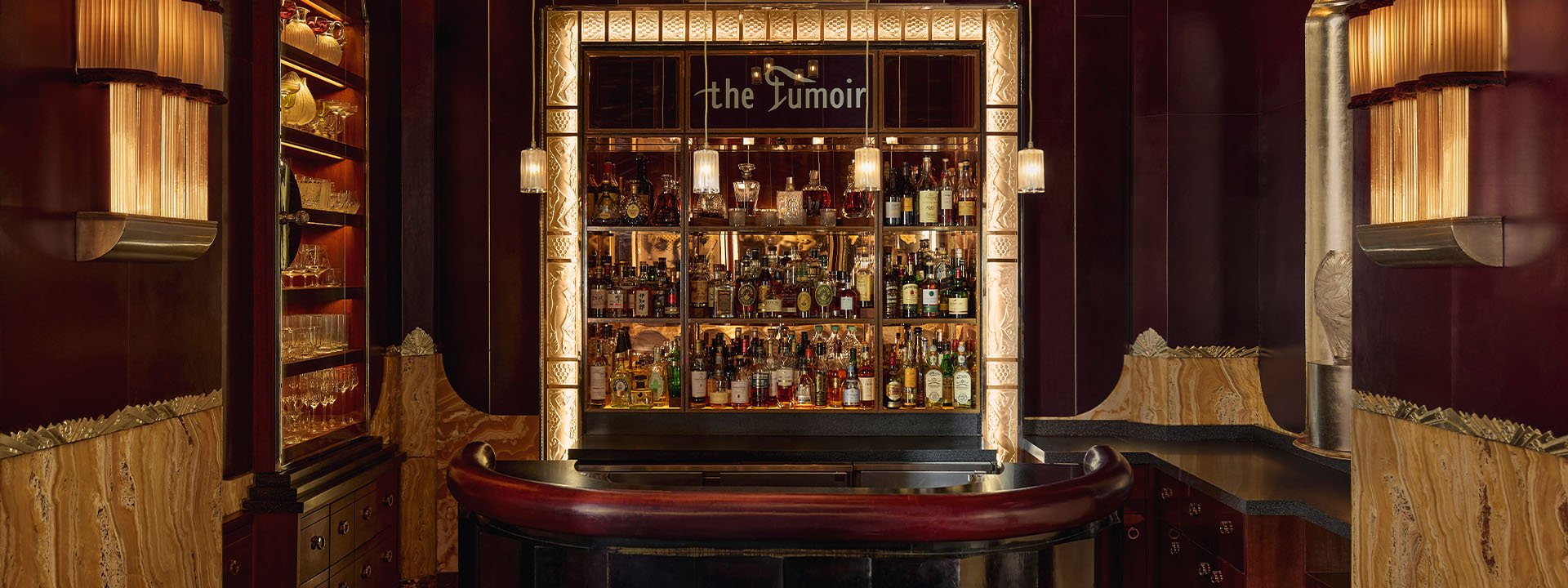 The dimly lit Fumoir bar at Claridge’s, showcasing a mirrored back bar stocked with spirits, art deco lighting, and rich maroon and gold interiors.