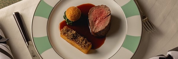 A fine dining dish featuring a medium-rare fillet of beef with rich sauce, layered potato, and a golden croquette atop wilted greens on an elegant plate.