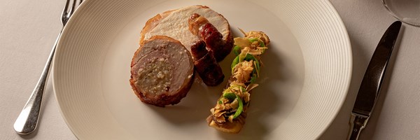 A plated festive dish of sliced roast turkey with bacon-wrapped sausage and a garnish of vegetables and crisp shavings arranged artfully on a white plate.