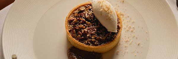 A dessert of pecan tart topped with a scoop of ice cream, served with a dollop of sauce and sprinkled with toasted crumbs on a white plate.