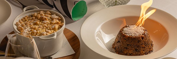 A festive dessert duo featuring a flaming Christmas pudding beside a pot of golden crumble dusted with icing sugar, presented on a white tablecloth.