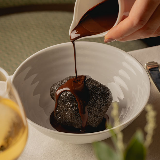Warm chocolate sauce being poured over a dark chocolate dome dessert in a white bowl.
