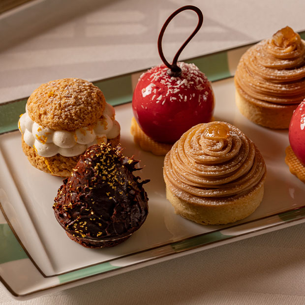 A selection of refined pastries on a rectangular plate, including glossy red domes, cream-filled choux, chocolate-covered confections, and tarts topped with caramel cream.