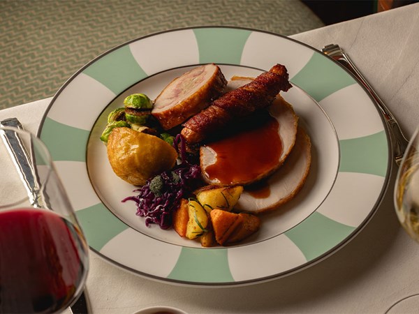 A plated Thanksgiving meal featuring sliced roast turkey with gravy, a pig in blanket, stuffing, roast potato, sautéed carrots, Brussels sprouts and red cabbage, served on a green-and-white striped Claridge’s plate with glasses of red and white wine in the foreground.