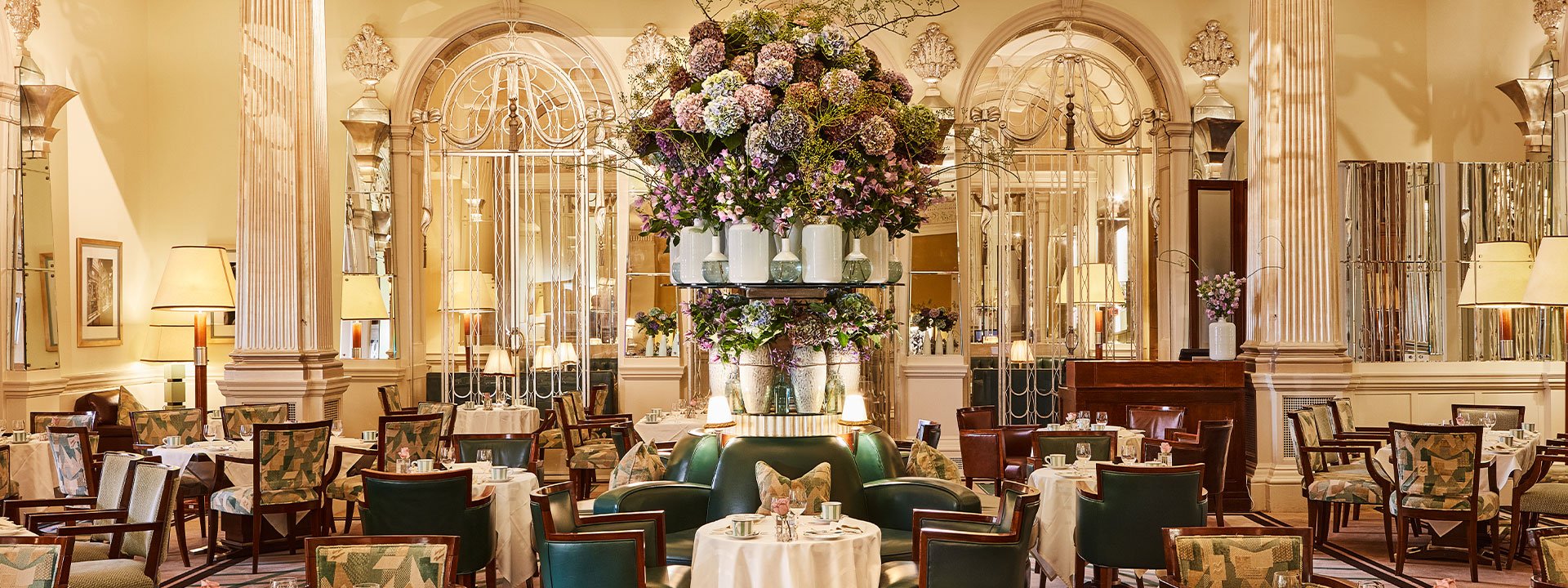 Elegant dining room at Claridge’s featuring high ceilings, ornate columns, mirrored panels, and a large central floral arrangement surrounded by set tables and armchairs.