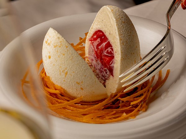 Intricate dessert resembling a cracked egg, filled with red fruit compote, resting in a nest of crisp pastry strands on a white plate.