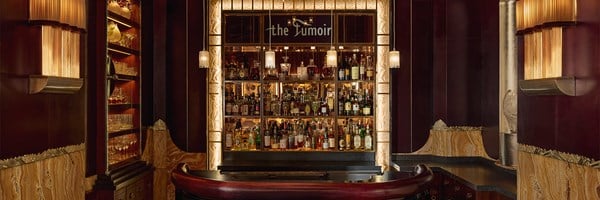Frontal view of The Fumoir bar at Claridge’s with three bar stools in front of a curved black and burgundy counter, illuminated glass shelves filled with spirits, and a monochrome portrait of a woman above the bar.