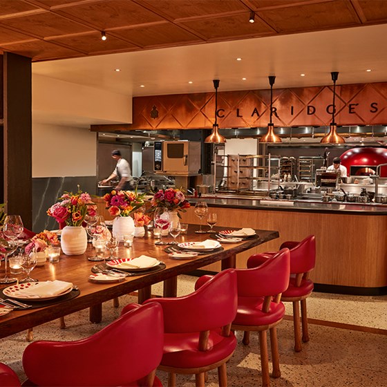 L’Epicerie’s intimate dining setup at Claridge’s, featuring a long wooden table surrounded by red leather chairs, elaborate table settings with candles and flowers, and an open kitchen with a striking red oven in the background.