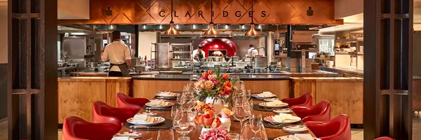 View of L’Epicerie at Claridge’s, showing an elegant private dining table set with floral centrepieces and polished tableware, facing an open kitchen with chefs at work beneath copper pendant lights and a copper canopy engraved with ‘Claridge’s’.