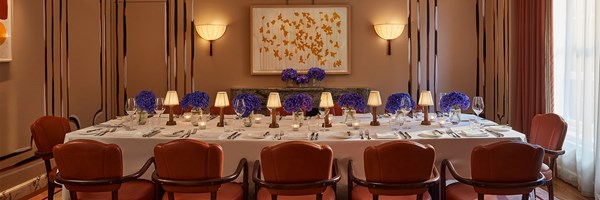 Claridge’s private dining room with a long table set in crisp white linen, surrounded by curved red chairs, small shaded table lamps, and vases of violet hydrangeas, with warm wall lights and modern artwork in a softly lit setting.
