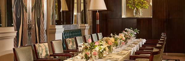 Elegant private dining space in Claridge’s Foyer and Reading Room, with a long banquet table adorned with pale pink and white floral arrangements, vintage-style upholstered chairs, art-deco mirrors, and a dark wood-panelled wall with modern lighting.