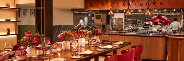 Private dining setting at L’Epicerie in Claridge’s, with a richly dressed wooden table featuring red leather chairs, floral centrepieces, and candlelight, set against an open kitchen with chefs working beneath copper pendant lights and a red domed oven.