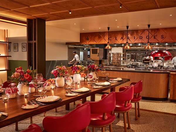 A vibrant chef’s table at Claridge’s, styled with red chairs, floral arrangements, and eclectic crockery, facing an open kitchen with copper lighting and active chefs.