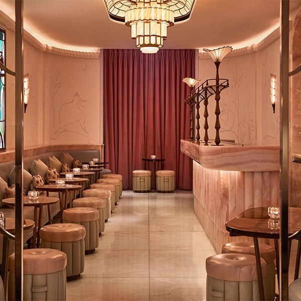 Wide view of The Painter’s Room at Claridge’s, showing a soft pink onyx bar with sculptural brass detailing, mirrored shelves with bottles and glassware, and a row of small tables and stools beside a banquette under a vibrant stained-glass window.