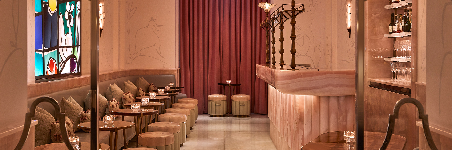 Wide view of The Painter’s Room at Claridge’s, showing a soft pink onyx bar with sculptural brass detailing, mirrored shelves with bottles and glassware, and a row of small tables and stools beside a banquette under a vibrant stained-glass window.