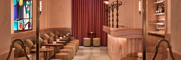 Wide view of The Painter’s Room at Claridge’s, showing a soft pink onyx bar with sculptural brass detailing, mirrored shelves with bottles and glassware, and a row of small tables and stools beside a banquette under a vibrant stained-glass window.