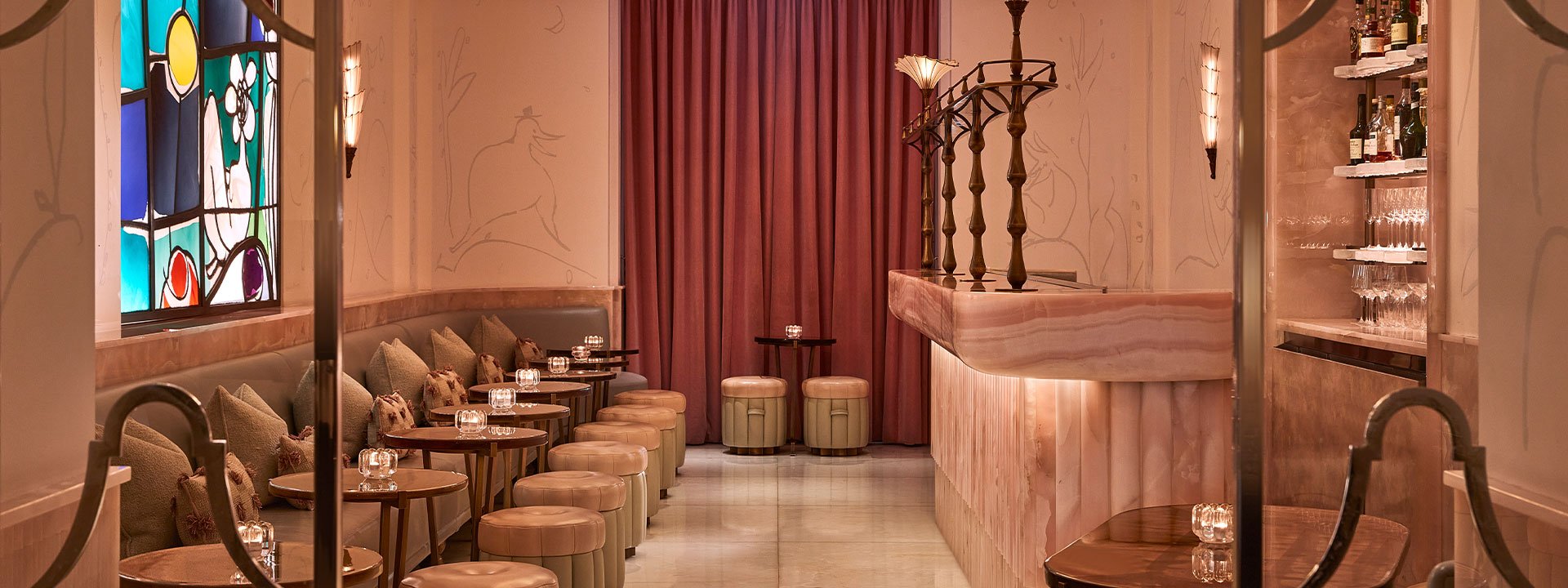 A softly lit Art Deco bar with pastel pink onyx counter, plush stools, and a stained glass window, evoking a refined and intimate atmosphere.