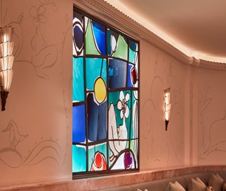 A vibrant stained-glass window with abstract shapes and bold colours, set in a softly lit blush-toned wall with line art illustrations and art deco sconces.