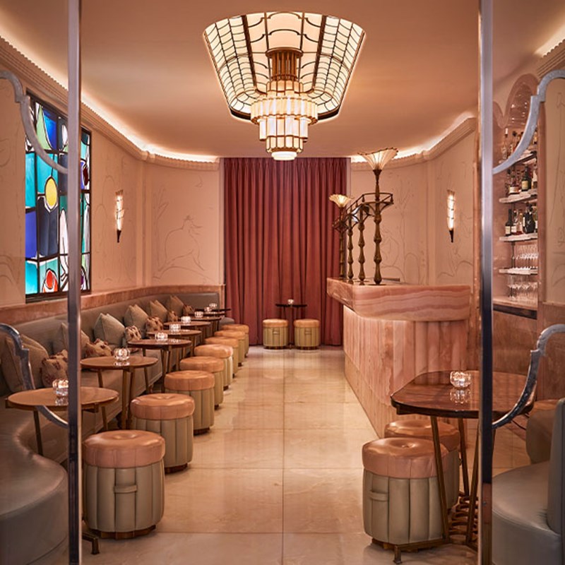 An elegant art deco bar room with soft pink and gold tones, featuring plush stools, marble flooring, and a sculptural chandelier hanging above a curved bar.