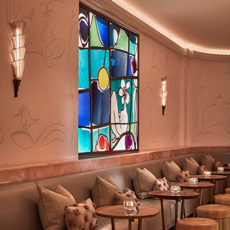A close-up of the Painter’s Room seating area with pale walls, scatter cushions, small round tables, and a colourful abstract stained-glass window.