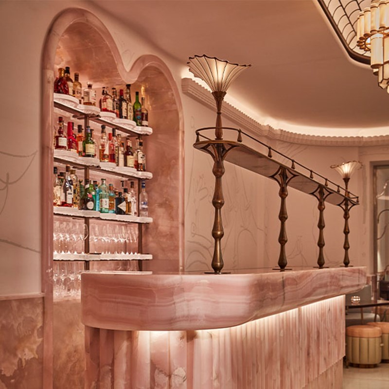 A view of the softly lit marble bar with a decorative bronze gantry and a backlit arched display of spirits and glassware.