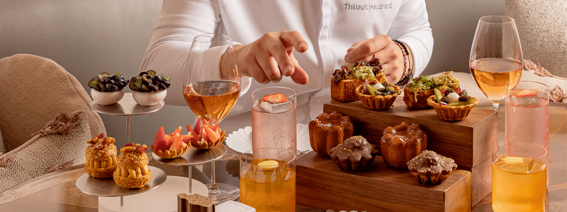 Close-up of an afternoon tea presentation at Claridge’s, with Chef Thibault Hauchard reaching toward a wooden stand of savoury tarts and pastries, surrounded by elegant desserts, fresh fruit, and sparkling drinks.