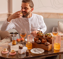 A smiling chef in a white uniform, seated at a marble table in a softly lit lounge, enjoys an elaborate afternoon tea spread with pastries, tarts, and drinks including rosé wine and cocktails.
