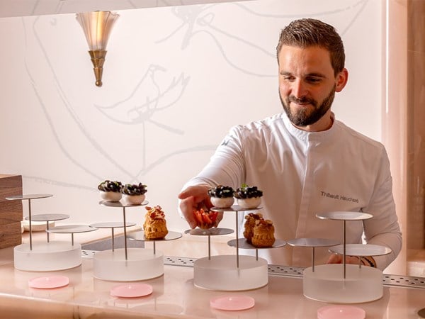 A pastry chef in a white uniform, named Thibault Hauchard, places colourful pastries on tiered white stands in an elegant setting with soft lighting and a wall illustration of birds.