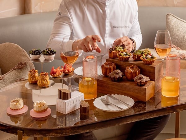 A chef seated at a marble table arranges an afternoon tea spread featuring a variety of artful pastries, chocolates, and drinks including pink and amber-toned beverages.