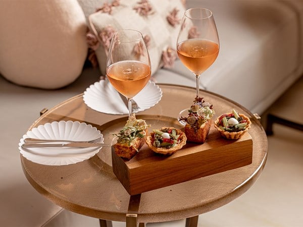 A close-up of a round table set for two with savoury tartlets and canapés on a wooden stand, flanked by two glasses of rosé wine and scalloped white plates with silver cutlery.