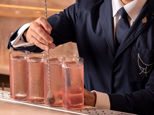 A bartender in a navy jacket and tie stirs one of several tall glasses of pink sparkling cocktails lined up on a bar, each with large square ice cubes.