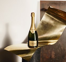 A bottle of Krug Grand Cuvée champagne elegantly rests on a curved sheet of golden metal, exuding a sense of luxury and refinement against a minimalist backdrop.