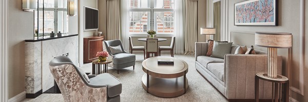 Claridges  Brook Suite living area. A window is to the back of the room and a circular wooden table, and chairs sit in front of it. A couch sits to the right and a round wooden table with a book sits in front. Two chairs sit next to the table.