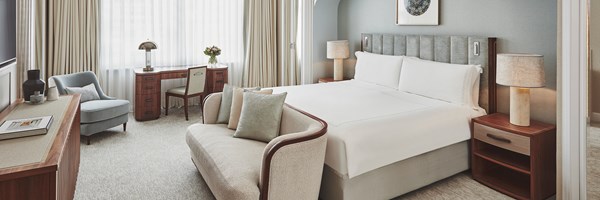 Claridges Brooke Suite bedroom. A bed sits to the right and two wooden bedside tables with a lamp sit beside it. A sofa sits at the end of the bed. A window is to the back of the room. A wooden desk with a lamp and vase with flowers sits in front of it.