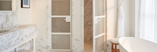 Light-filled marble bathroom featuring a freestanding white bathtub, walk-in shower, and a robe hanging beside tall sheer-curtained windows.