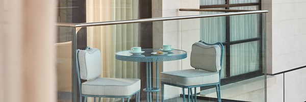 Compact glass-fronted balcony with a mint-blue bistro set for two, set with tea and biscuits, overlooking the hotel’s cream façade.