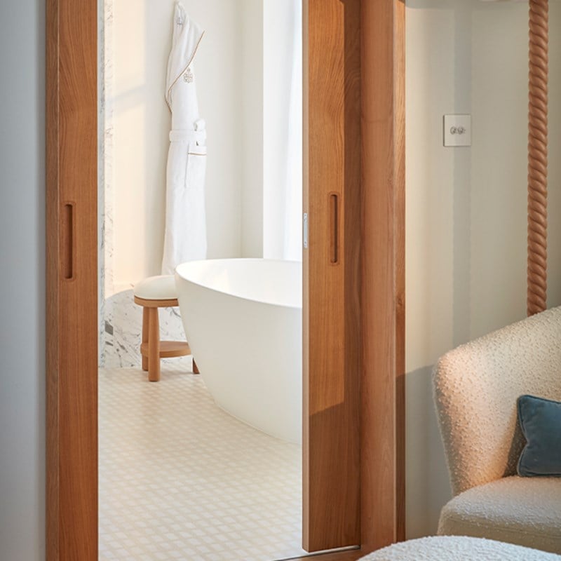 Partial view into a bright bathroom with freestanding tub and hanging robe, seen through wooden sliding doors from the bedroom.