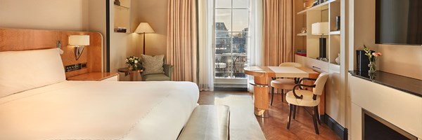 View of a serene guest room featuring a large bed, armchair with side table and flowers, and a private balcony seen through floor-to-ceiling windows.