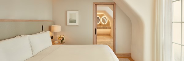 Peaceful bedroom scene with a large bed, bedside lamp and flowers, and a softly lit bathroom visible through a wooden-framed door.