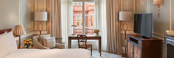 Classic guestroom interior with a writing desk by the window, armchair, and view of red brick architecture outside.
