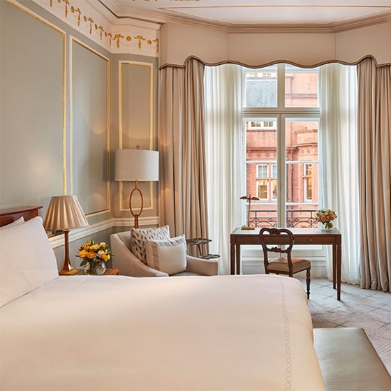 Elegant Claridge’s Room featuring a king-size bed, classic moulded walls with gold detailing, a sitting area with armchair and floor lamp, and a writing desk beside a large window overlooking red brick architecture.