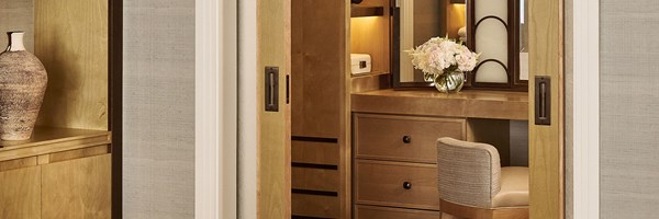 Refined dressing area with sliding wooden doors revealing a vanity table, upholstered chair, illuminated mirror, and a glass vase of pale flowers, with built-in cabinetry and textured wall covering.
