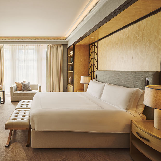 Elegant studio bedroom with a king-sized bed dressed in white linens, a cushioned bench at the foot, soft beige and gold tones throughout, and a sofa positioned by floor-to-ceiling windows with sheer curtains.