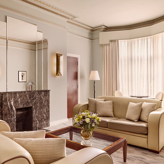 Spacious suite living room with cream art deco sofas, a dark marble fireplace, mirrored wall panels, and a bay window with sheer curtains.