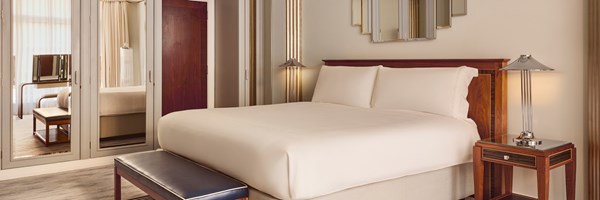 Wide-angle view of the suite bedroom featuring a king bed with white linens, mirrored wardrobe doors, a dark wood bench, and chrome lamps.