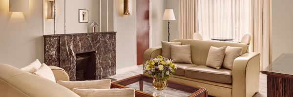 Panoramic view of the suite’s living room with curved cream sofas, a glass coffee table, a marble fireplace, and large windows with sheer curtains.