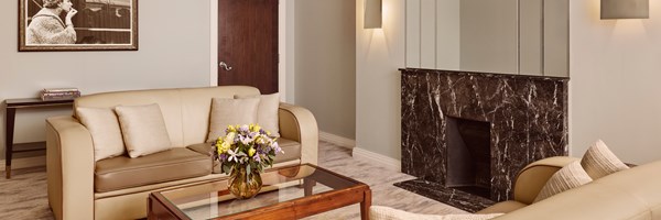 Wide-angle of the living room highlighting cream sofas, a glass table with flowers, a dark marble fireplace, and a framed black-and-white photograph.