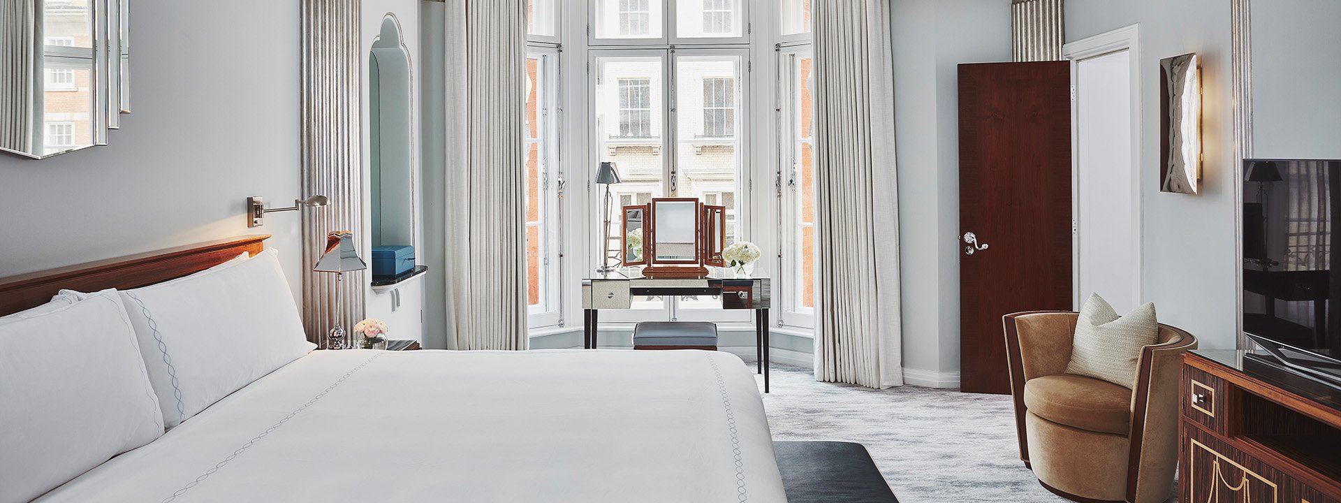 A view of the bed in a bedroom from Claridge's Suite, with a dressing table and lots of natural daylight in the interior.